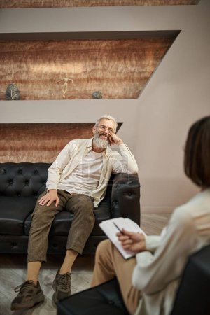glücklicher bärtiger älterer Mann mittleren Alters mit Tätowierung sitzt auf Ledercouch und schaut zum Psychologen