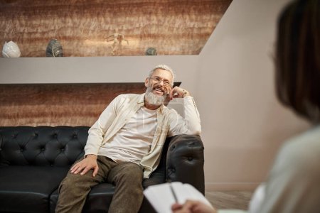 cheerful bearded middled aged man with tattoo sitting on leather couch and looking at psychologist