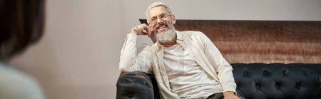 Téléchargez les photos : Joyeux barbu middled homme âgé assis sur le canapé en cuir et en regardant psychologue, bannière - en image libre de droit