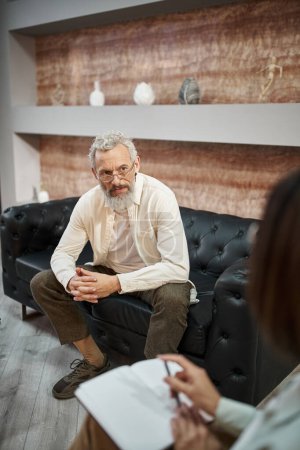 bearded middled aged man with tattoo sitting on leather couch and listening to female psychologist