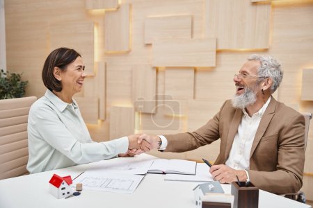 Foto de Mujer feliz sonriendo y estrechando las manos con agente inmobiliario de mediana edad en la oficina de bienes raíces, venta de propiedades - Imagen libre de derechos