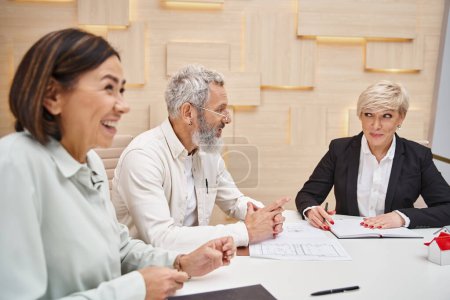 happy middle aged man smiling near wife and female realtor while sitting near blueprint of new home