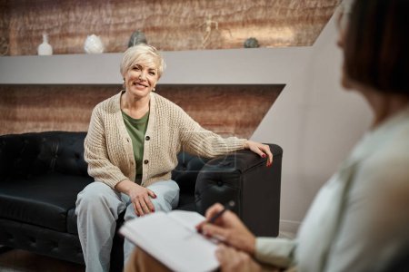 cheerful blonde middled aged woman sitting on leather couch and looking at psychologist on session