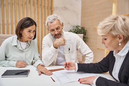 cheerful middle aged couple smiling near female realtor and looking at blueprint of new home