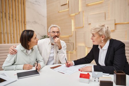cheerful middle aged married couple smiling near female realtor and discussing project of new house