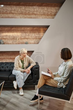 blonde middled aged woman sitting on couch and talking to female psychologist during therapy session