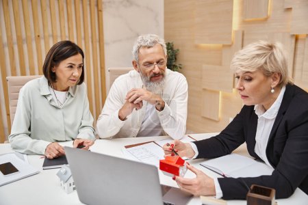 middle aged realtor showing carton house model to married couple in real estate office, property