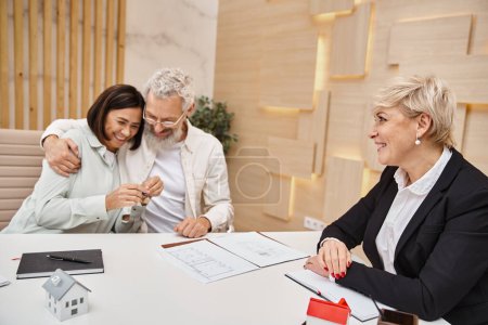 cheerful married couple holding key from new house near smiling realtor in real estate office