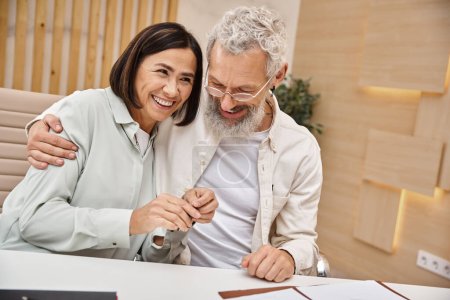 happy bearded man hugging multiracial wife holding key from new house in real estate office