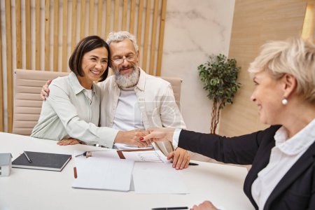 Foto de Mujer feliz estrechando la mano con agente inmobiliario cerca de marido y haciendo trato en la oficina de bienes raíces - Imagen libre de derechos