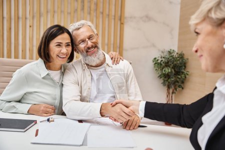 Foto de Hombre feliz de mediana edad estrechando la mano con el agente inmobiliario cerca de la esposa y haciendo trato en la oficina de bienes raíces - Imagen libre de derechos