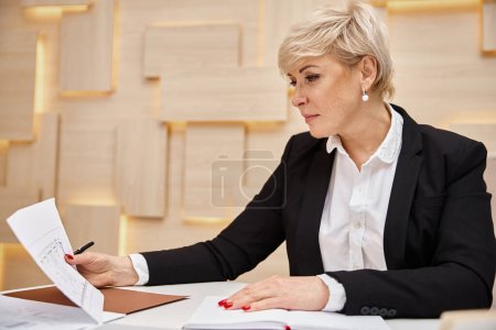 blonde middle aged realtor with short hair working in real estate office, looking at blueprint