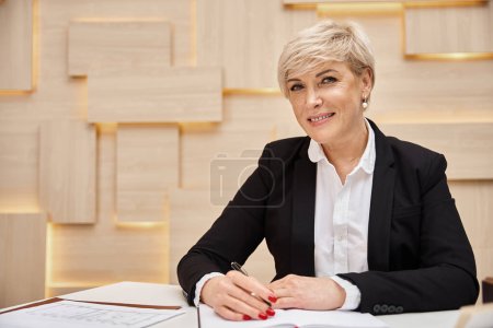 happy blonde middle aged realtor with short hair working in real estate office, looking at camera