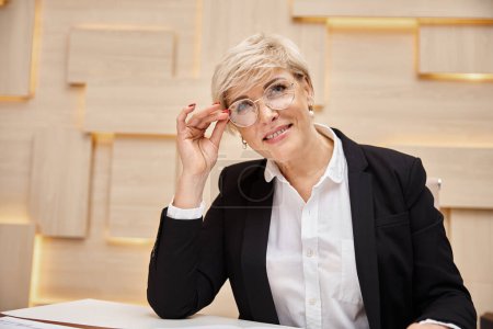 glückliche Blondine mittleren Alters Makler mit Brille, die im Immobilienbüro arbeitet, schaut in die Kamera