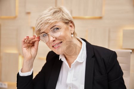 happy blonde middle aged realtor in eyeglasses working in real estate office, looking at camera