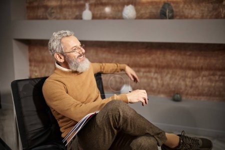 happy middle aged psychologist in glasses smiling and sitting on armchair during therapy session