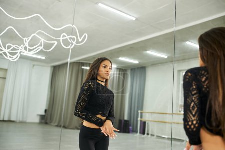 künstlerische afrikanisch-amerikanische Tänzerin in schwarzer Kleidung, die in den Spiegel schaut und im Tanzsaal trainiert