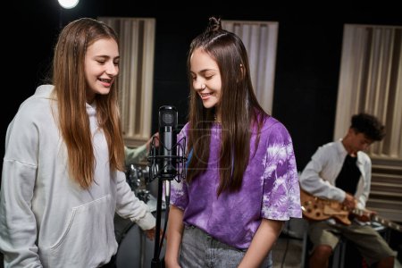 Photo for Cheerful adorable teenage girls singing while their friends playing drums and guitar, musical group - Royalty Free Image