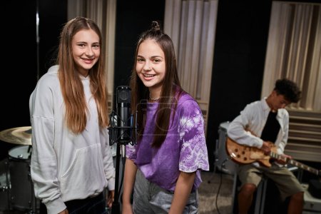 Foto de Alegre bonita adolescente niñas mirando a la cámara cerca de micrófono con su amigo tocando la guitarra - Imagen libre de derechos