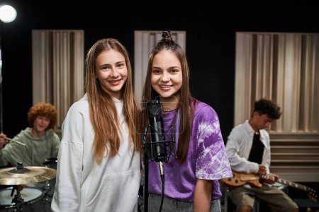 fröhliche Teenager-Mädchen, die in die Kamera neben dem Mikrofon schauen, während ihre Freunde Schlagzeug und Gitarre spielen