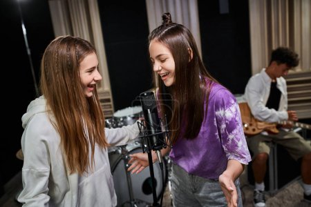 cheerful pretty teenage girls in casual attire singing with friend playing guitar on backdrop