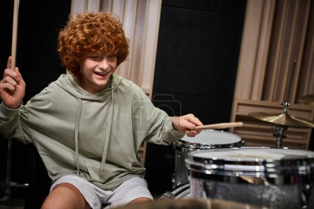 alegre adorable pelirrojo adolescente en traje casual tocando la batería activamente mientras en el estudio