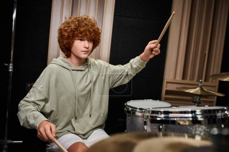 adorable adolescente concentrado con pelo rojo en traje de cada día tocando su batería mientras está en el estudio
