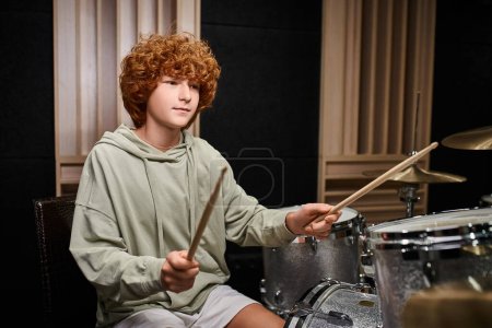 adorable centrado adolescente con el pelo rojo en traje acogedor casual tocando su batería en el estudio