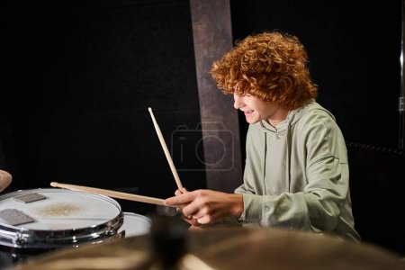 fröhlicher talentierter Teenager mit roten Haaren in lässiger bequemer Kleidung, der im Studio Schlagzeug spielt