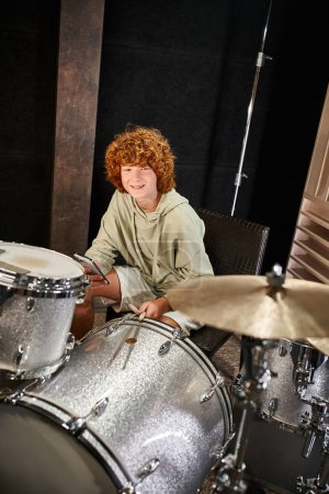 cheerful adorable red haired teenage boy in cozy attire holding mobile phone next to his drums