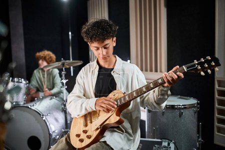 focus on cute talented teenage boy playing guitar with his blurred drummer on backdrop in studio