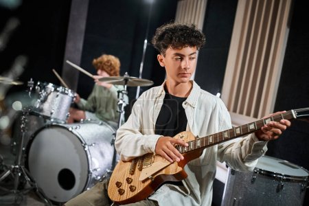 Konzentrierter entzückender Teenager in lässiger Kleidung, der neben seinem verschwommenen Schlagzeuger Gitarre spielt