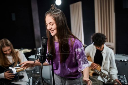 Téléchargez les photos : Joyeuse brune adolescente chantant joyeusement avec ses amis jouant des instruments en studio - en image libre de droit
