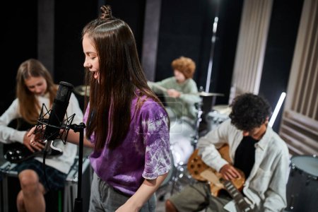 Téléchargez les photos : Joyeux adorables adolescents en tenue décontractée vibrante chanter et jouer des instruments en studio - en image libre de droit