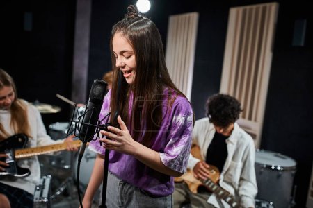 focus on cute teenage girl in vivid attire singing next to her blurred band members in studio