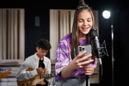 lustiges entzückendes Teenager-Mädchen, das neben ihrem verschwommenen Gitarristen singt und auf das Smartphone schaut