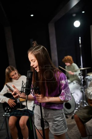 cheerful teenage girl in vivid attire singing and looking at her mobile phone next to her friends