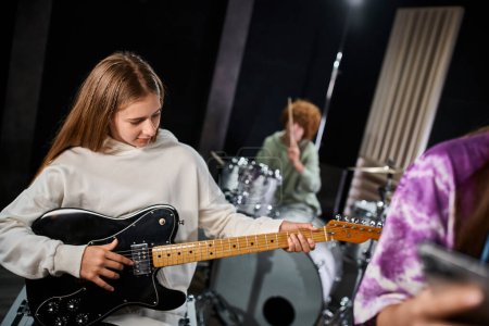 adorable rubia adolescente en traje casual tocando la guitarra junto a sus talentosos amigos en el estudio