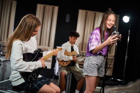 Foto de Alegre linda adolescente en traje diario cantando felizmente al lado de su amigo en guitarras en estudio - Imagen libre de derechos