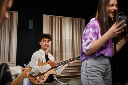 Foto de Adorable lindo adolescente tocando la guitarra y mirando atentamente a sus amigos, grupo musical - Imagen libre de derechos