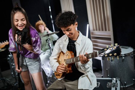 Téléchargez les photos : Joyeuse adolescente mignonne chantant à côté de son ami mignon jouant de la guitare et un autre garçon à la batterie - en image libre de droit