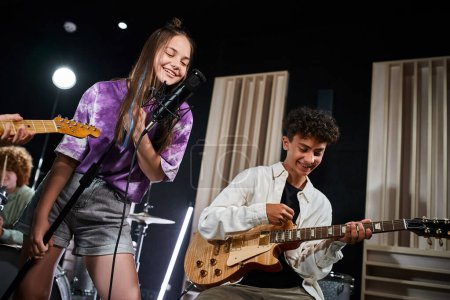Téléchargez les photos : Adorable adolescente joyeuse chantant joyeusement tandis que son ami jouant des instruments en studio - en image libre de droit