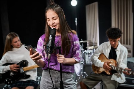 Foto de Se centran en la alegre adolescente en traje vívido cantando y mirando el teléfono cerca de sus guitarristas borrosos - Imagen libre de derechos