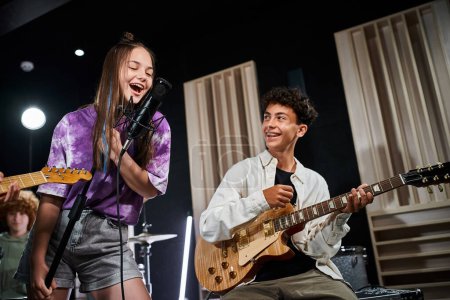 Téléchargez les photos : Adolescents talentueux gais en tenues décontractées chantant et jouant de la guitare alors qu'ils sont dans leur studio - en image libre de droit