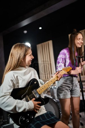 Photo pour Se concentrer sur une adolescente blonde joyeuse jouant de la guitare et regardant son amie brune chanter - image libre de droit