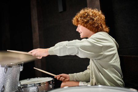 fröhlich entzückender rothaariger Teenager in lässigem, gemütlichem Outfit, der aktiv im Studio Schlagzeug spielt