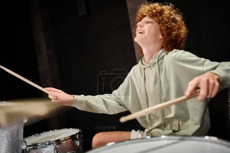fröhlicher talentierter rothaariger Teenager in bequemer Alltagskleidung, der im Studio Schlagzeug spielt