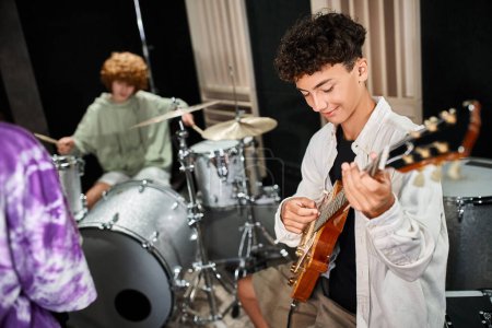focus on cheerful cute teenage boy playing guitar actively next to his blurred band members