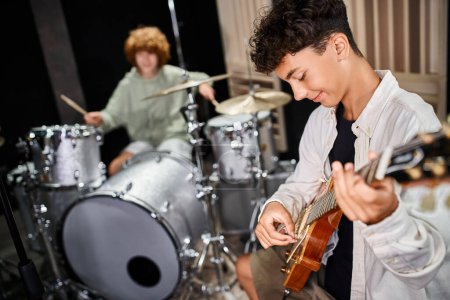 focus on talented adorable teenage boy playing guitar actively next to his blurred drummer in studio