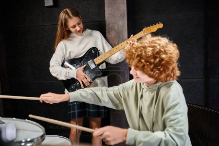 Fokus auf fröhliches hübsches Teenager-Mädchen, das neben ihrem verschwommenen rothaarigen Schlagzeuger ihre Gitarre spielt